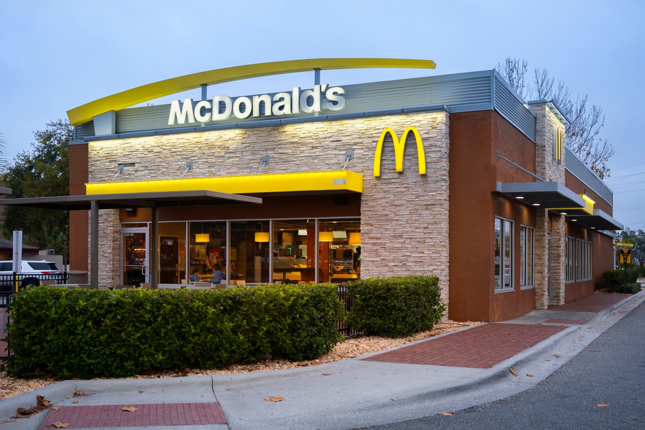A McDonald’s restaurant with a yellow arch logo and illuminated sign, surrounded by bushes and outdoor seating, during early evening with lights on inside.
