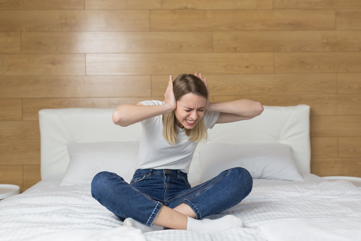 A woman sits cross-legged on a bed, covering her ears with her hands and closing her eyes tightly, appearing frustrated or bothered, with a wooden wall in the background.