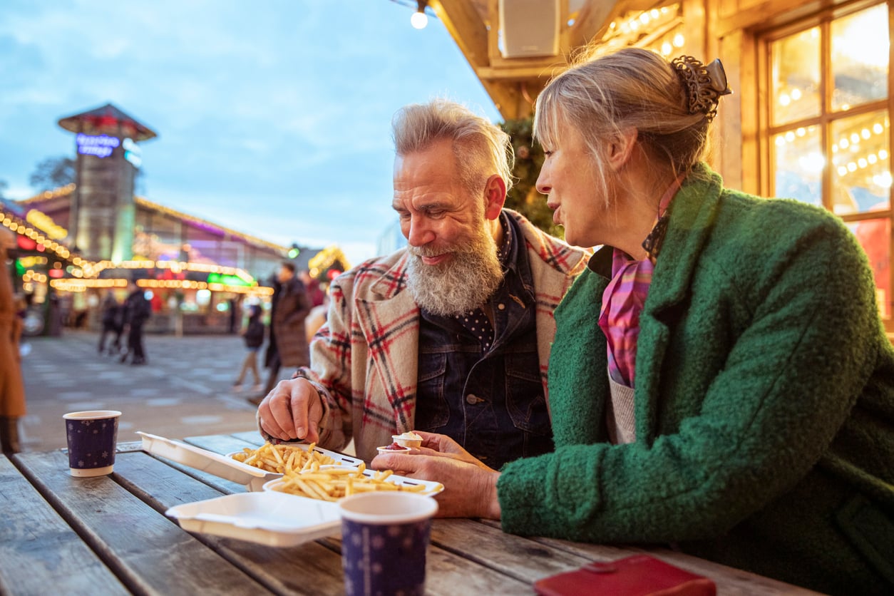 An older couple sits at an outdoor table, sharing fries and smiling at each other, with festive lights and a fairground in the background—a perfect snapshot of unforgettable vacation experiences.