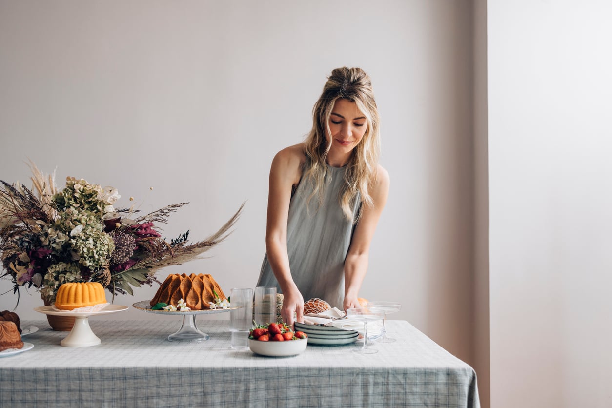 A woman in a gray dress arranges desserts on a table set with cakes, strawberries, plates, and a floral arrangement against a plain, light-colored wall.