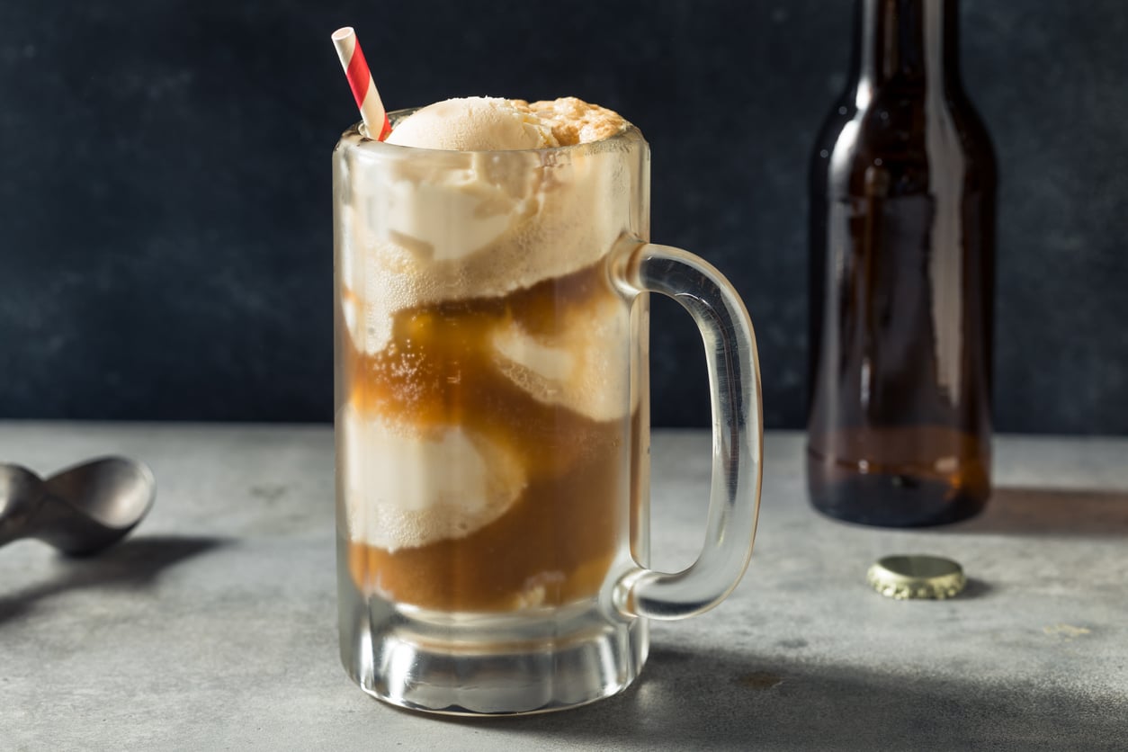 A frosty mug of root beer float with scoops of vanilla ice cream and a striped straw, sits on a gray surface beside a brown bottle and bottle cap.