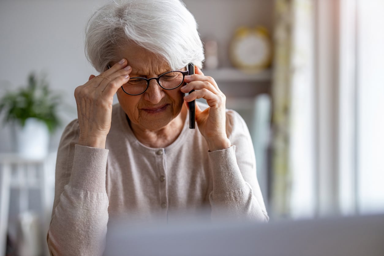 Elderly woman with white hair and glasses holds her head in pain while talking on the phone, sitting indoors with a concerned expression.