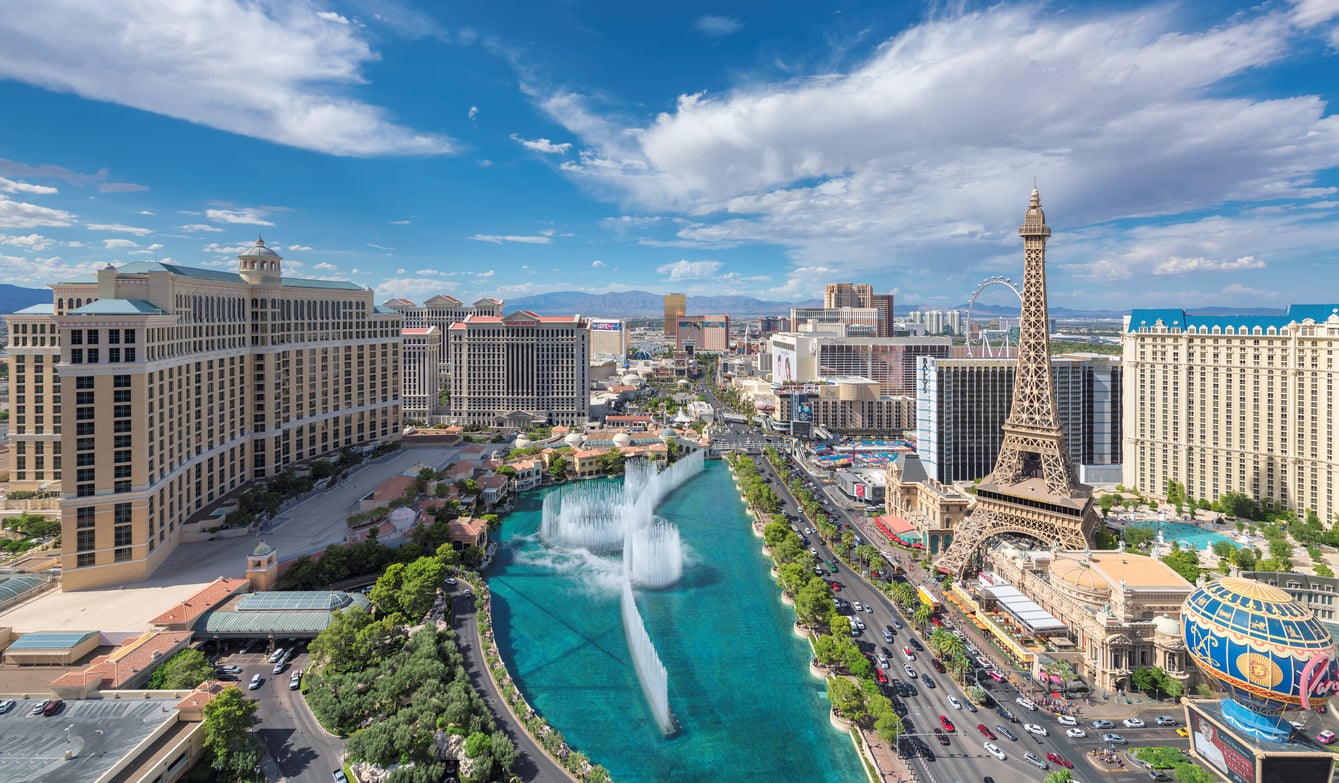 Aerial view of Las Vegas Strip featuring the Bellagio fountains, Paris Las Vegas’ Eiffel Tower replica, and bustling casinos under a bright blue sky with scattered clouds.