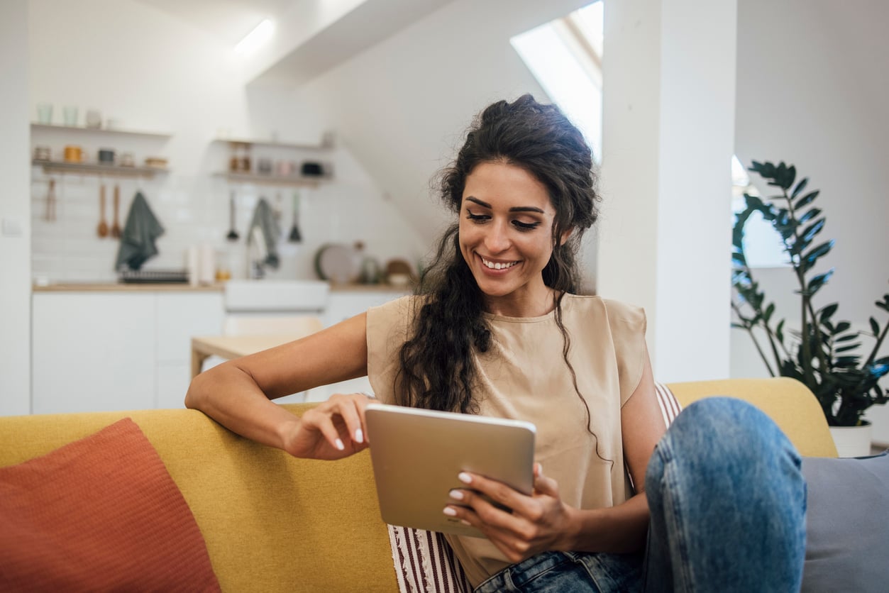 A woman sits on a yellow couch in a modern living room, smiling while using a tablet. The background shows a kitchen with shelves and plants.