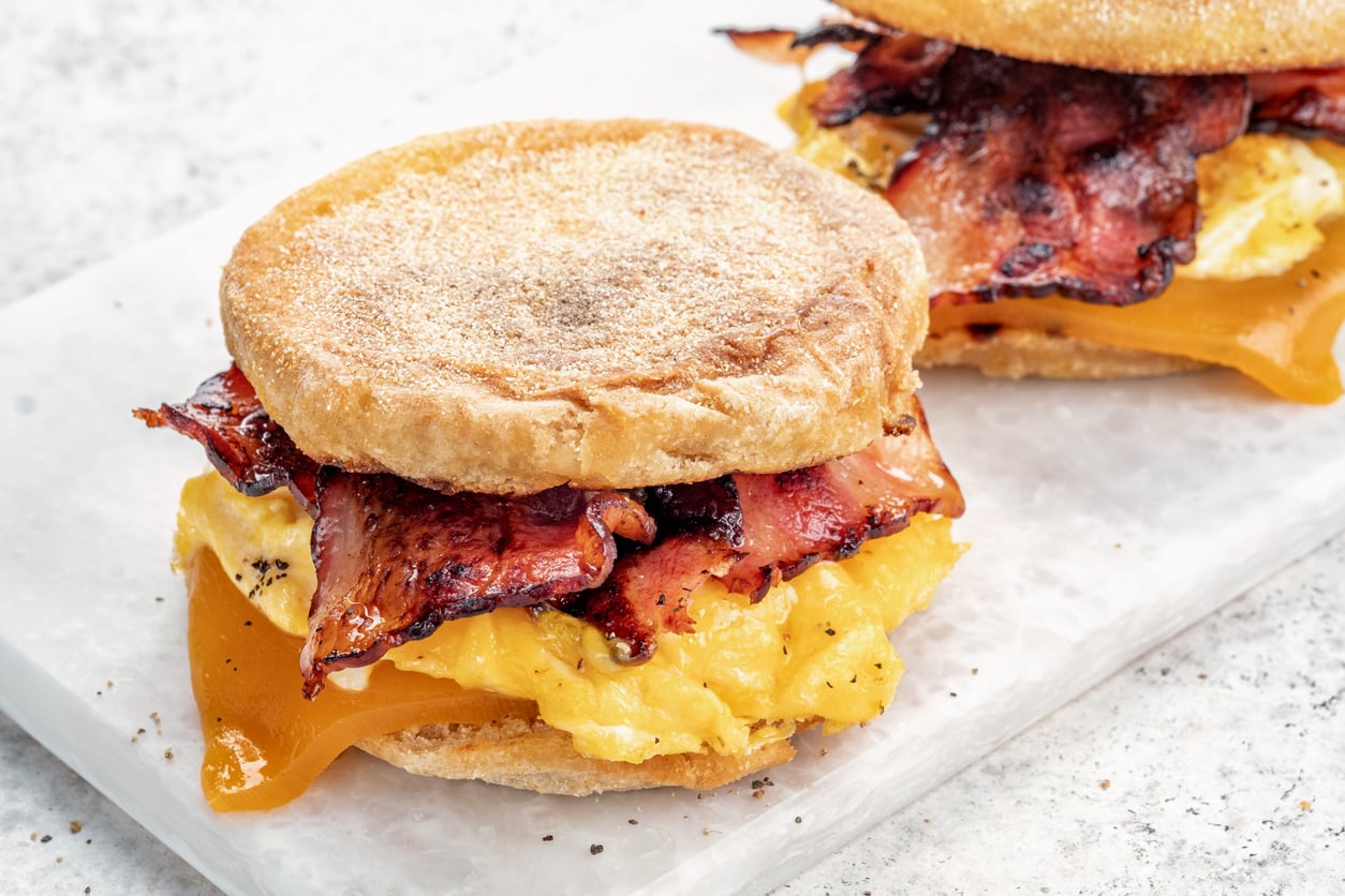 An English muffin breakfast sandwich with scrambled eggs, crispy bacon, and cheddar cheese on a white marble surface. Another sandwich is partially visible in the background.
