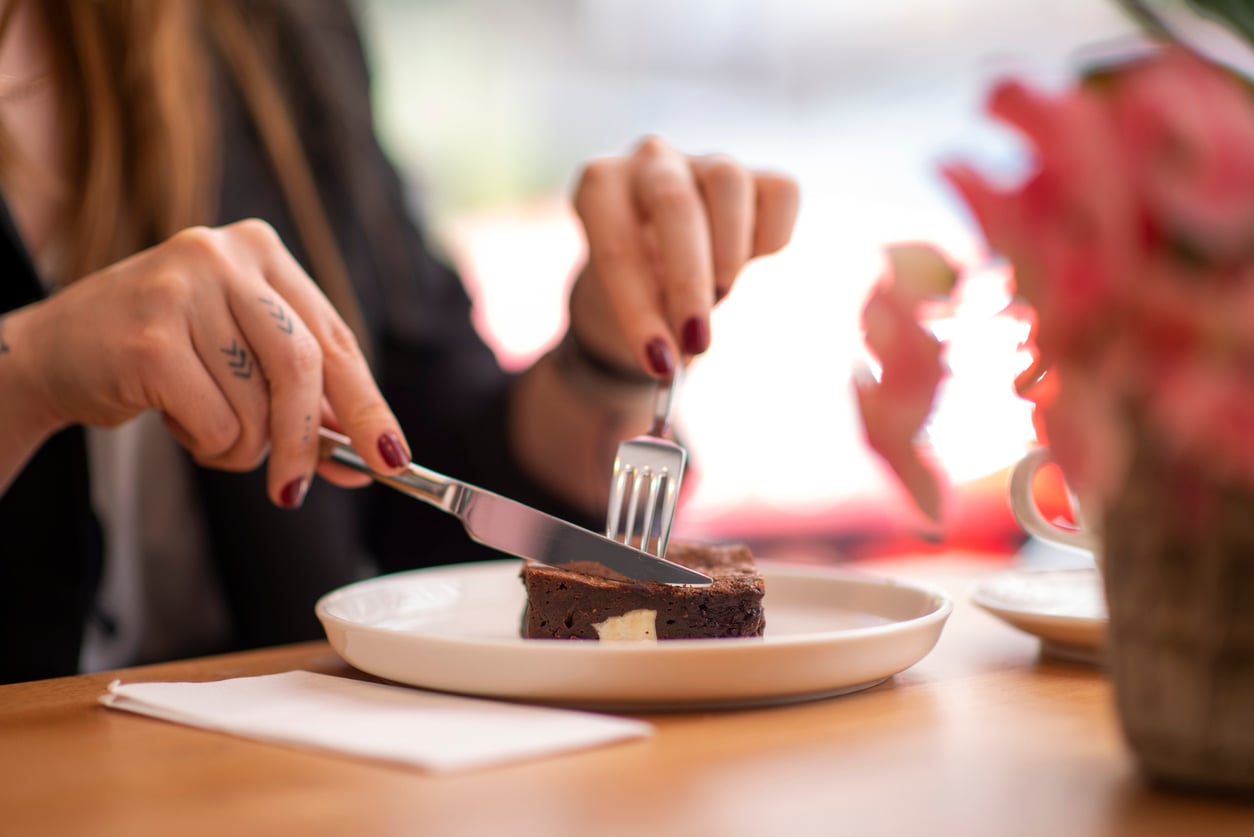 A person with painted nails uses a fork and knife to cut into a brownie on a white plate, with a pink flower and napkin on the table nearby.