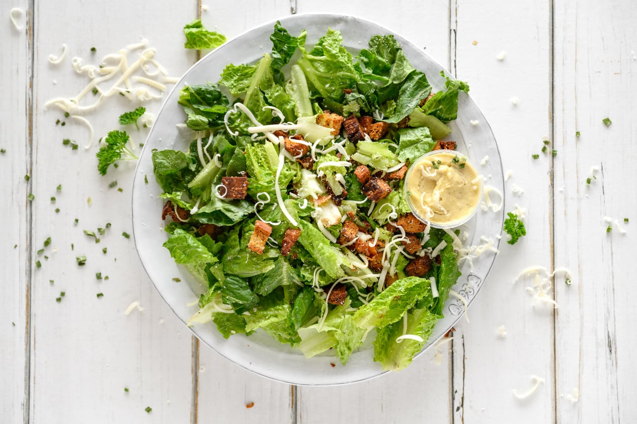 A plate of Caesar salad with romaine lettuce, croutons, shredded cheese, and a small cup of creamy dressing on the side, all on a white wooden table with scattered cheese and herbs.