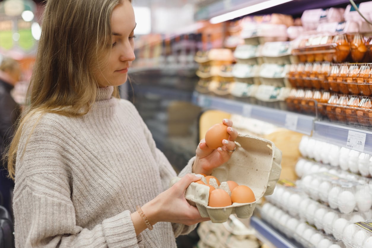A woman wearing a beige sweater holds an open carton of eggs, examining them while shopping in a grocery store egg aisle.