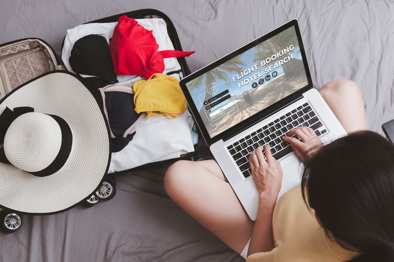 A woman sits by an open suitcase with clothes and a hat, using a laptop for flight booking and hotel search, preparing for a trip.