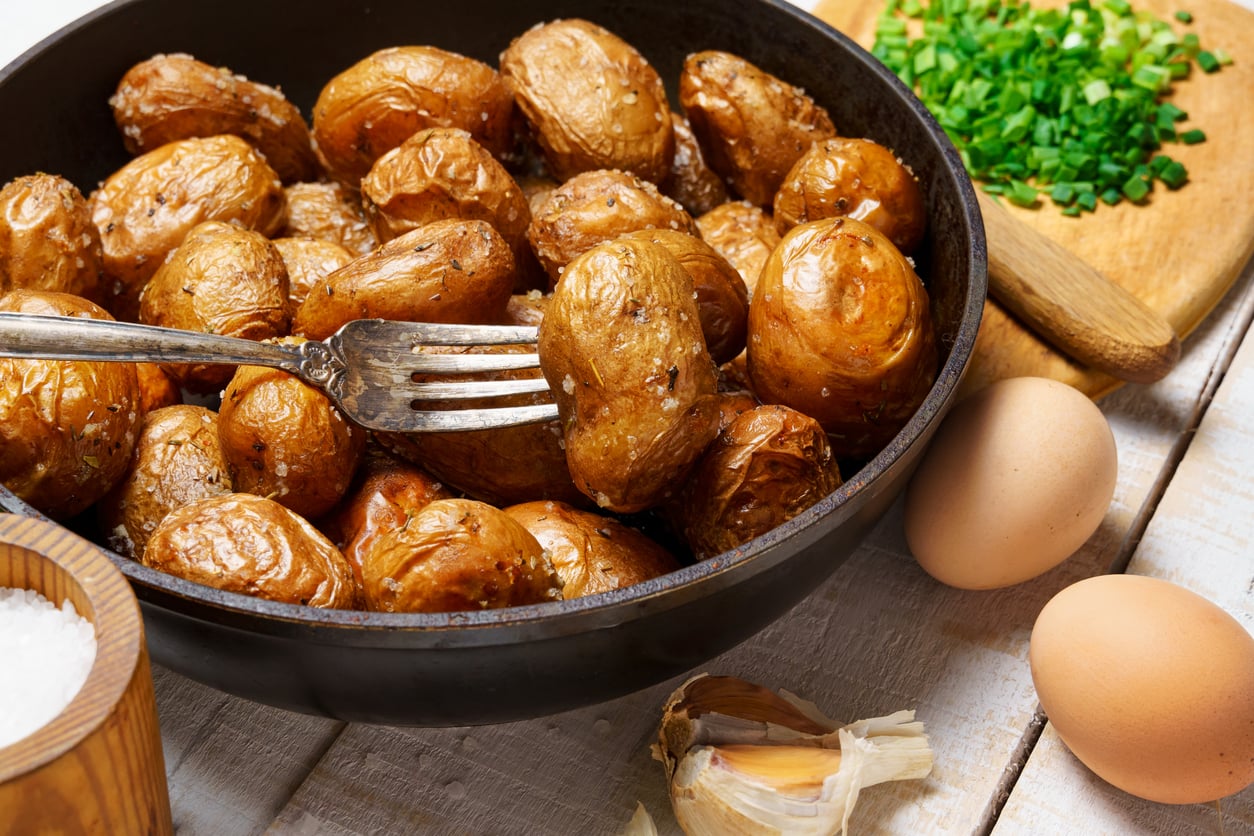 A fork holds a roasted potato in a pan filled with roasted baby potatoes, reminiscent of comforting restaurant specialties. Nearby are eggs, chopped chives on a cutting board, a bowl of salt, and a garlic clove on a rustic white table.