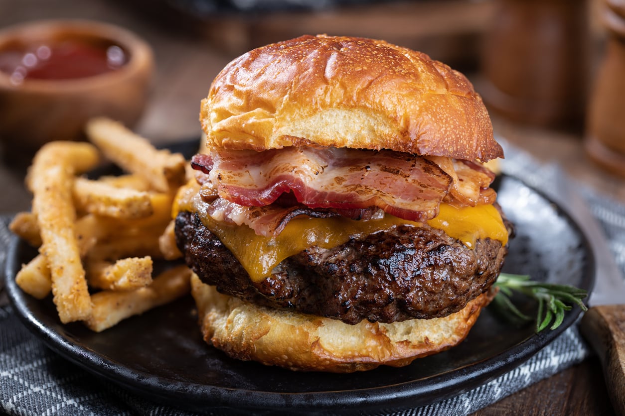 A close-up of a cheeseburger with bacon on a brioche bun, served with seasoned French fries on a dark plate. A small bowl of ketchup is blurred in the background.