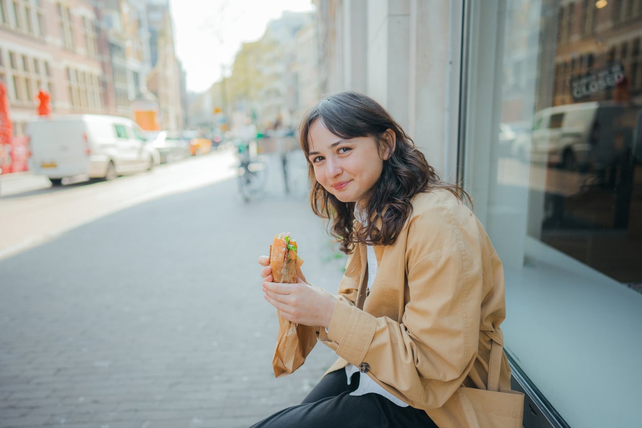 Woman in a tan coat sits on a city sidewalk, smiling and holding a sandwich. Buildings, a street, and bicycles are visible in the background on a sunny day.