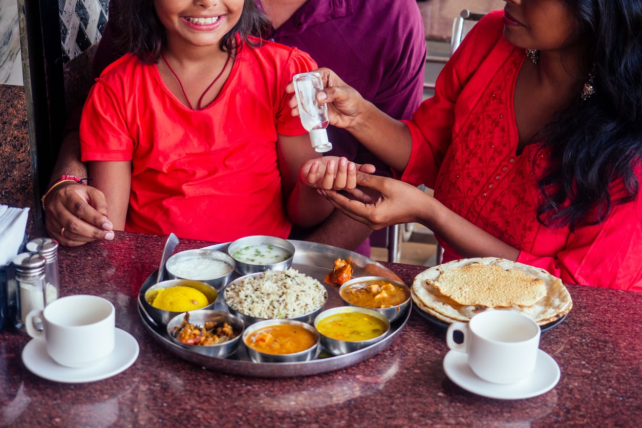 A woman applies hand sanitizer to a smiling child’s hand at a table set with Indian food, including rice, curries, yogurt, and papad, with two cups of tea or coffee.