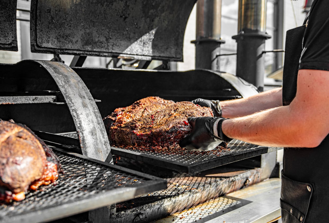A person wearing black gloves places a large smoked brisket onto a metal rack inside a commercial barbecue smoker, preparing one of the top foods to order at any true barbecue joint.