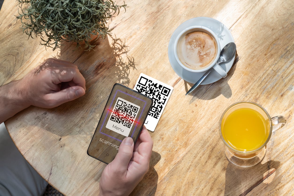 A person scans a QR code labeled “Menu” on a table with a smartphone. There is a cup of coffee, a glass of orange juice, and a small plant on the wooden table.