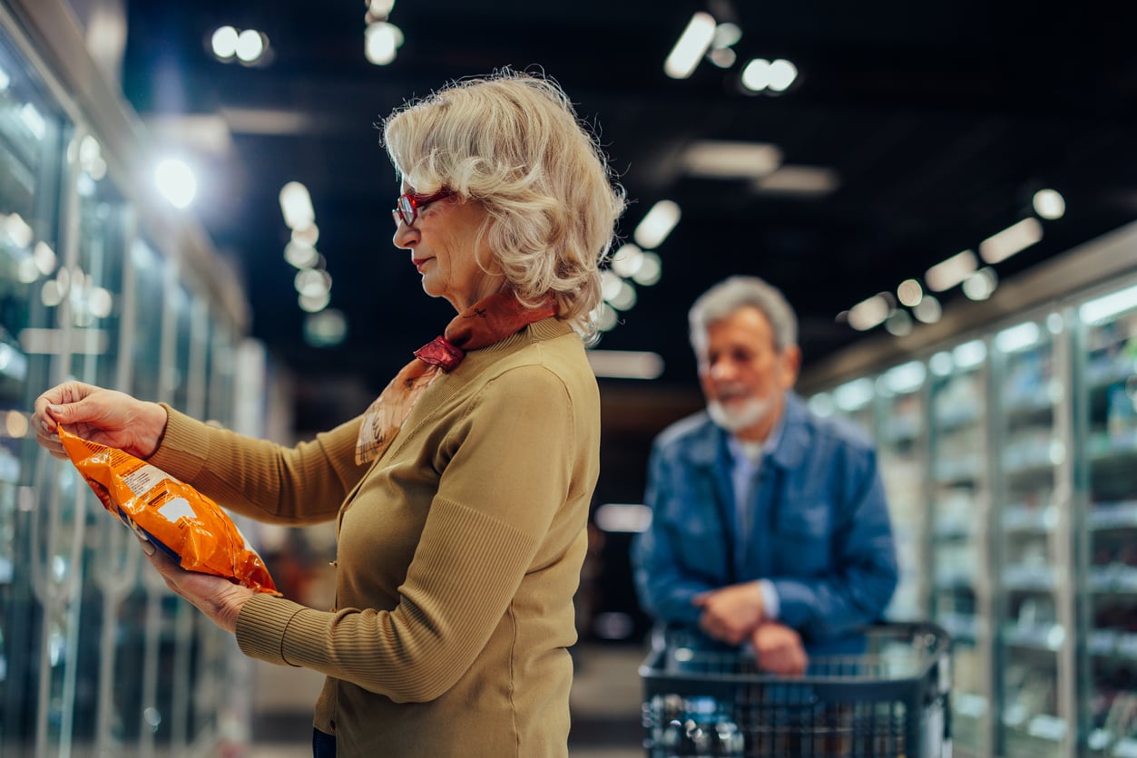 An older woman reads a package label in a grocery store aisle while an older man stands behind her with a shopping cart.