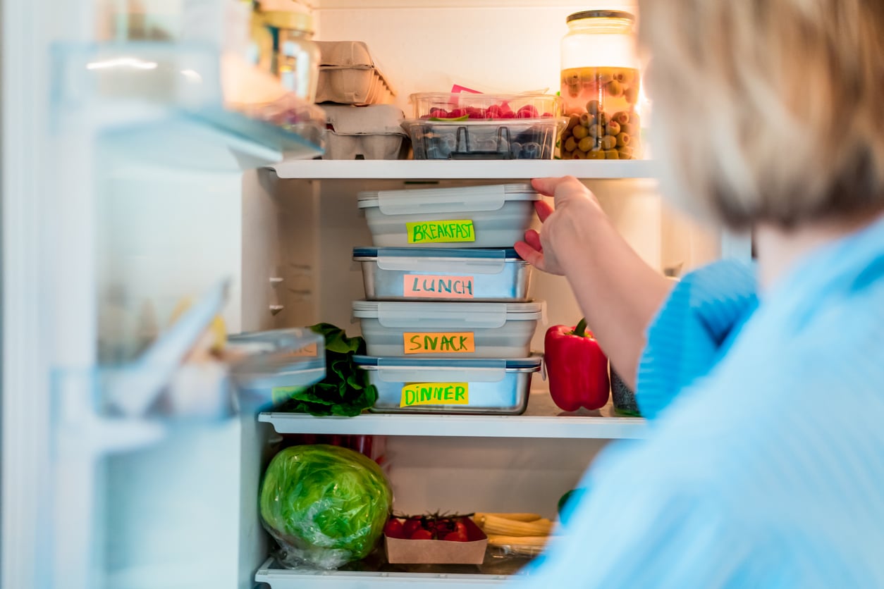 A person reaches into a refrigerator stocked with labeled food containers reading “BREAKFAST,” “LUNCH,” “SNACK,” and “DINNER,” along with fresh vegetables and jars.