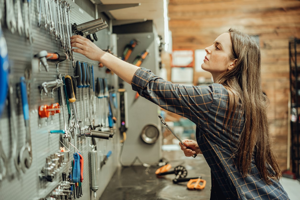 A woman in a plaid shirt reaches for a tool on a wall-mounted pegboard in a workshop, holding a wrench in her other hand. Various tools are organized on the pegboard and workbench.
