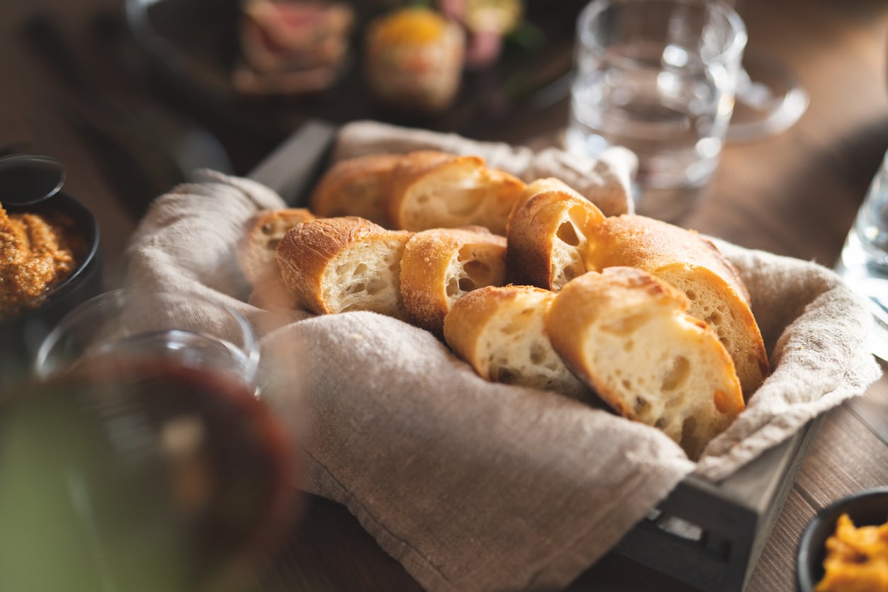 A basket lined with a beige cloth holds several slices of crusty baguette bread on a wooden table, with blurred dishes and glasses in the background.