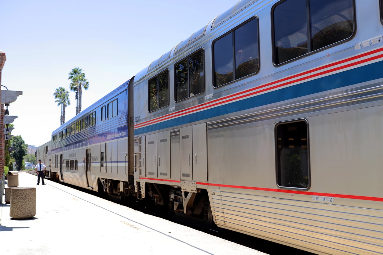 A silver double-decker passenger train with blue and red stripes is stopped at a sunny station platform. A conductor stands nearby, and palm trees are visible in the background.