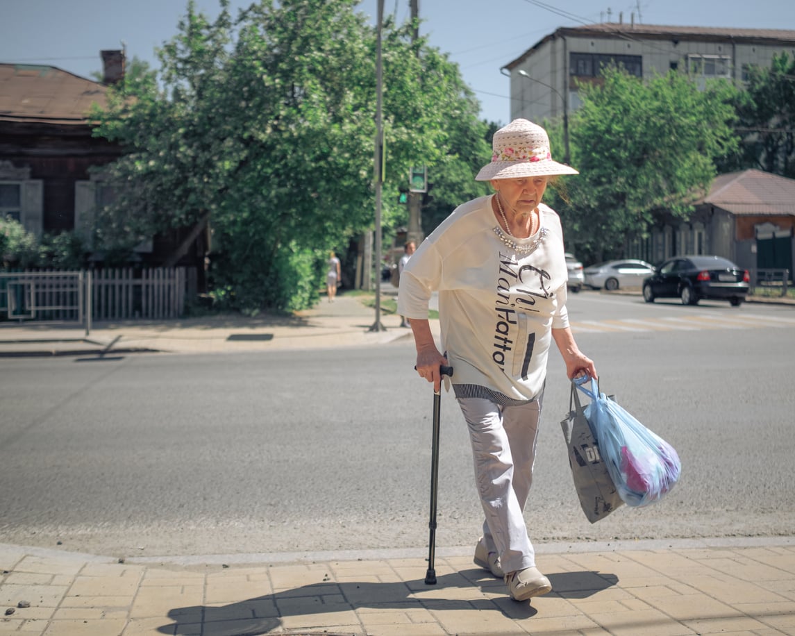 Elderly woman wearing a sunhat walks with a cane on a sunny day, carrying plastic bags, crossing a quiet street with houses and parked cars in the background.