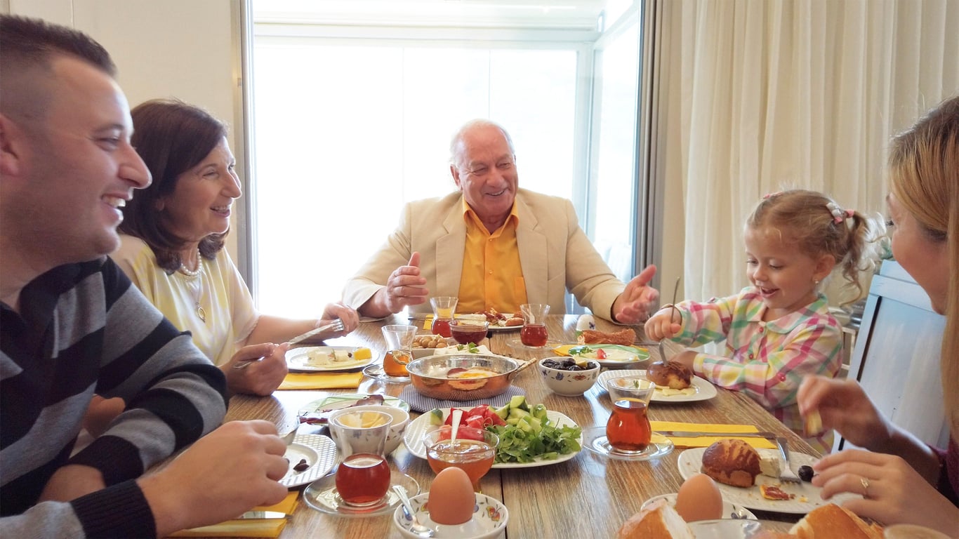 Five people of different ages are smiling and enjoying a meal together at a dining table filled with various foods, including eggs, bread, salads, and tea in glass cups. A young girl is laughing with the others.