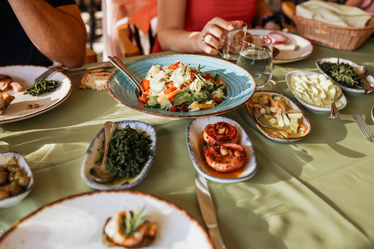 A table set with a green tablecloth features various dishes, including a salad, grilled tomatoes, greens, and bread. A person in a red top sits at the table, reaching for a glass of water.