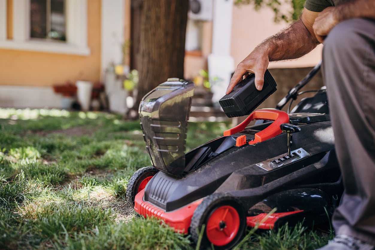 A person inserts a battery into a cordless lawn mower on a grassy yard near a house, preparing it for use.