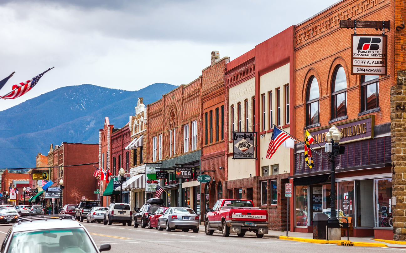 A small-town street with brick storefronts, American flags, parked cars, and mountains in the background under a cloudy sky.