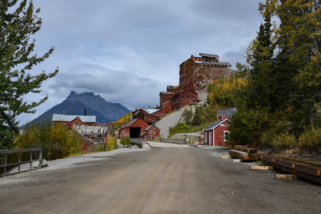 A dirt road leads to red historic mill buildings on a hillside, surrounded by autumn trees and mountains under a cloudy sky.