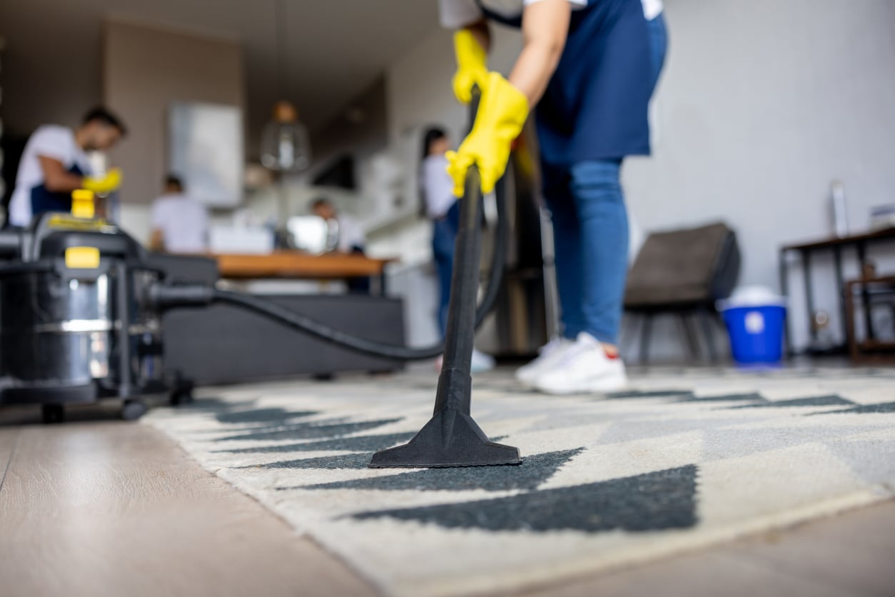 A person wearing yellow gloves vacuums a patterned rug in a living room, with other people cleaning in the background.