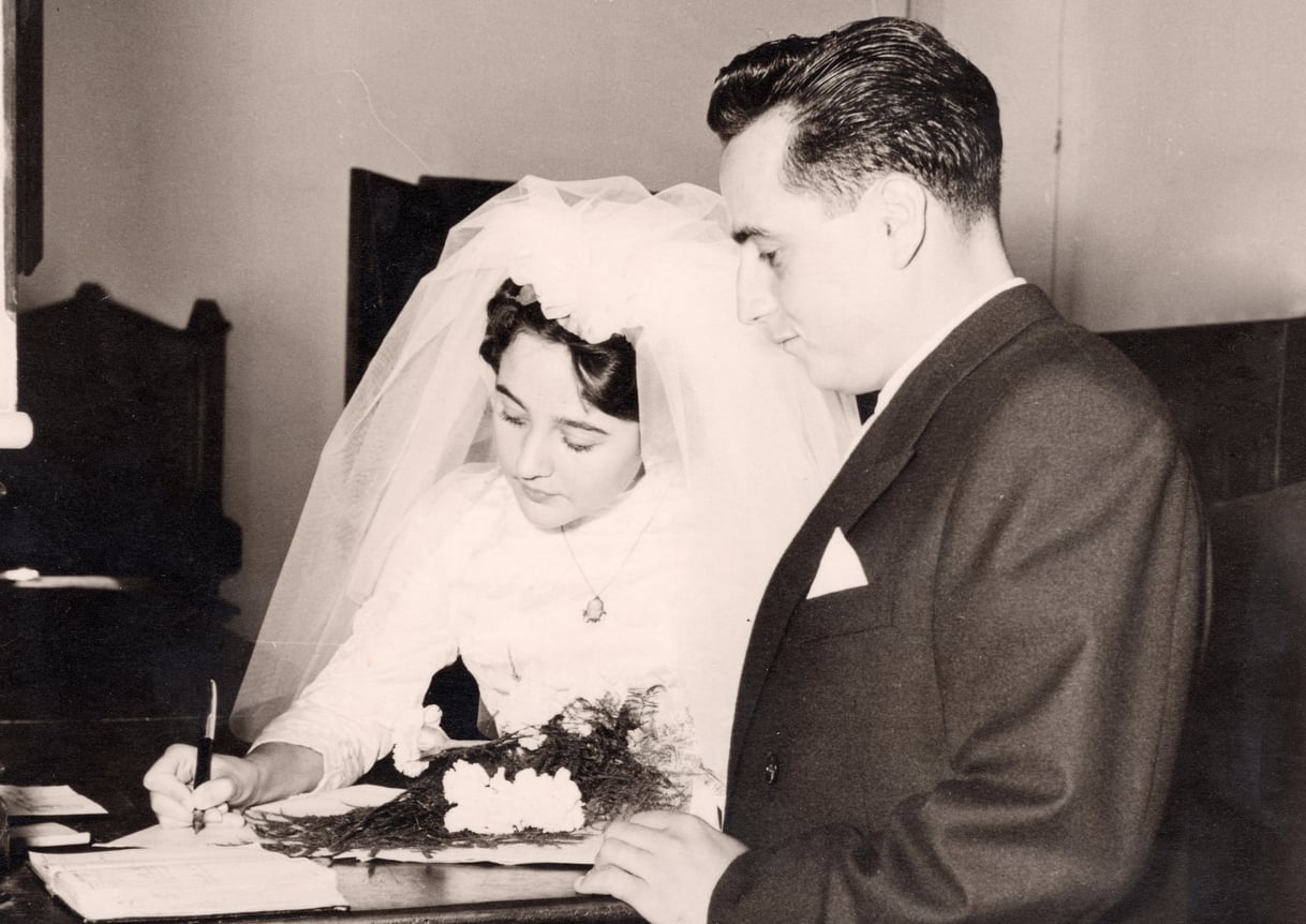 A bride in a white dress and veil signs a document while a groom in a suit stands beside her. They are at a table with papers and flowers, likely during a wedding ceremony.