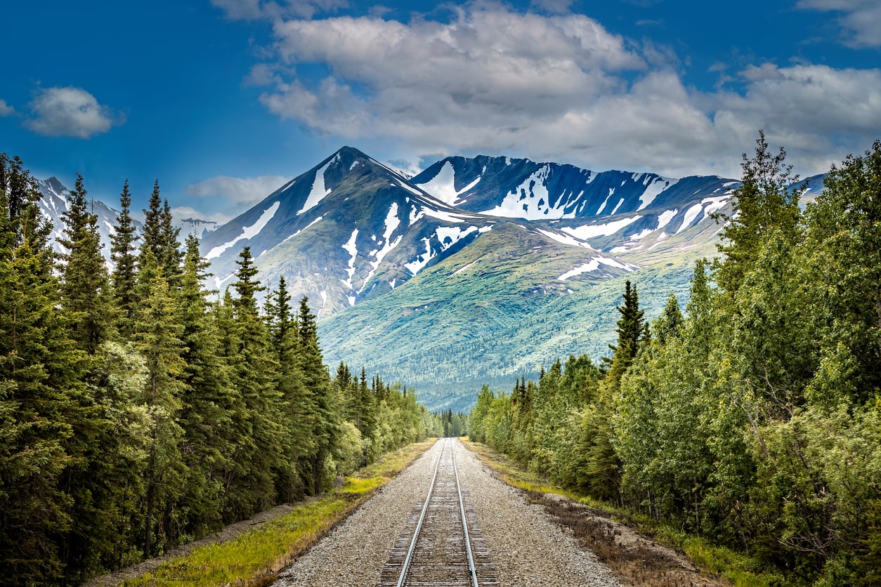 Railroad tracks run through a dense forest toward tall, snow-capped mountains under a partly cloudy blue sky.