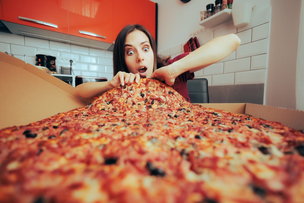 A surprised woman sits in a kitchen, holding a gigantic pizza that covers the table in front of her, with wide eyes and an excited expression.