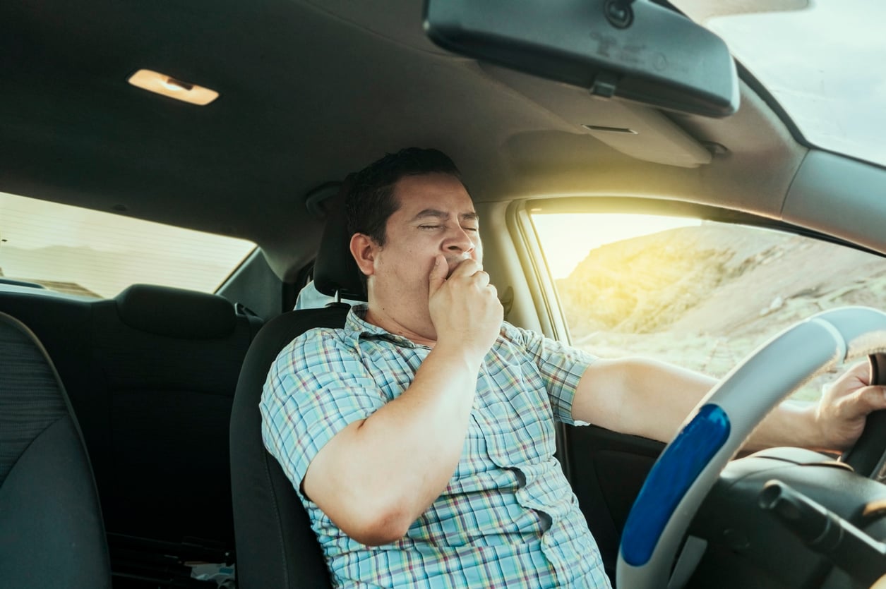 A man in a plaid shirt yawns while driving a car, covering his mouth with one hand. Sunlight shines through the window, and a mountainous landscape is visible outside.