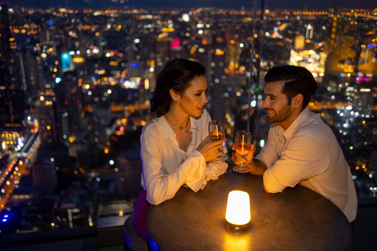 A couple sits at a rooftop table at night, sharing drinks and conversation. City lights glow in the background, and a small candle flickers on the table between them.