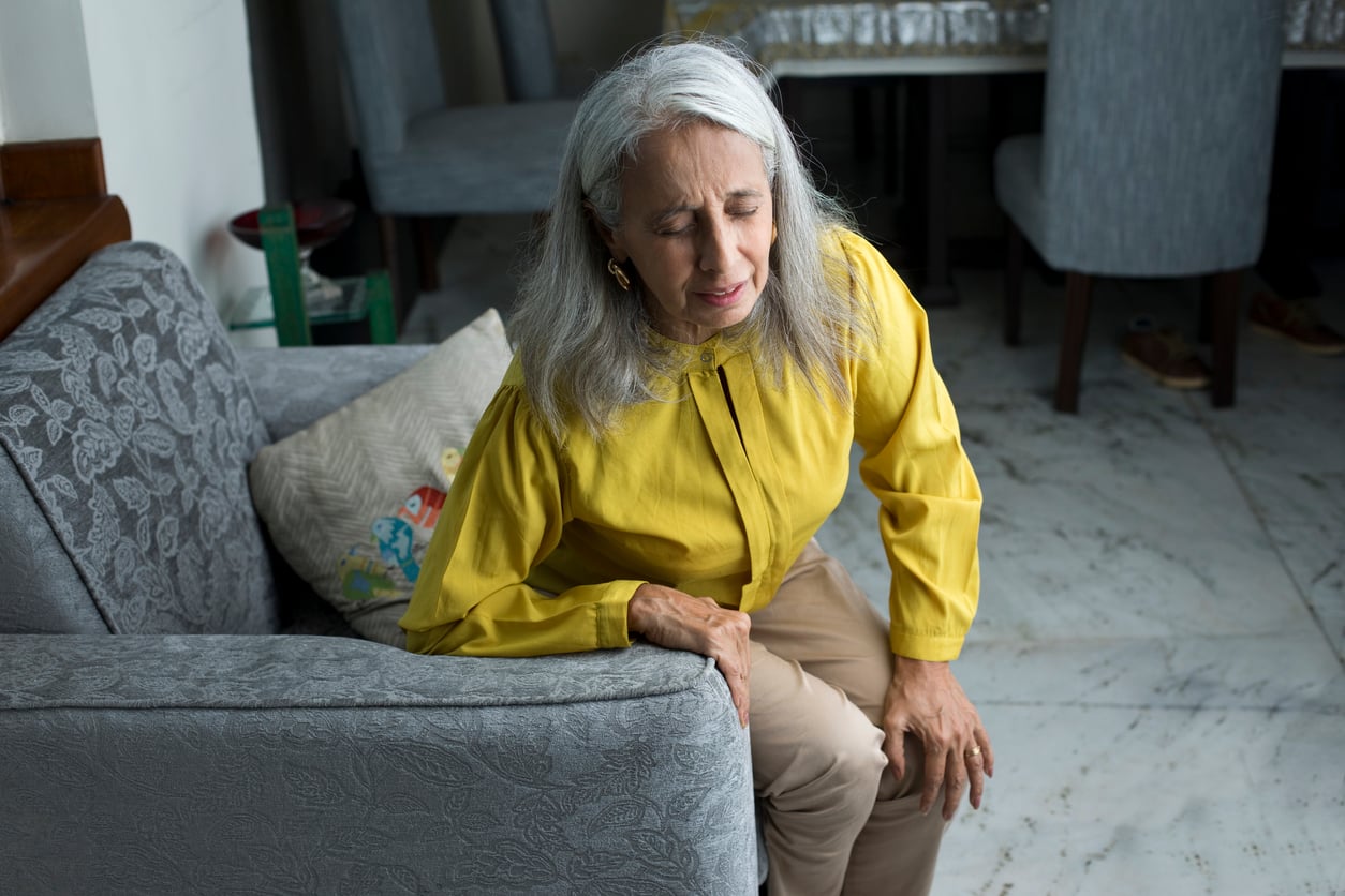 An older woman with gray hair, wearing a yellow blouse, sits on a sofa and holds her knee with a pained expression, suggesting discomfort or knee pain.