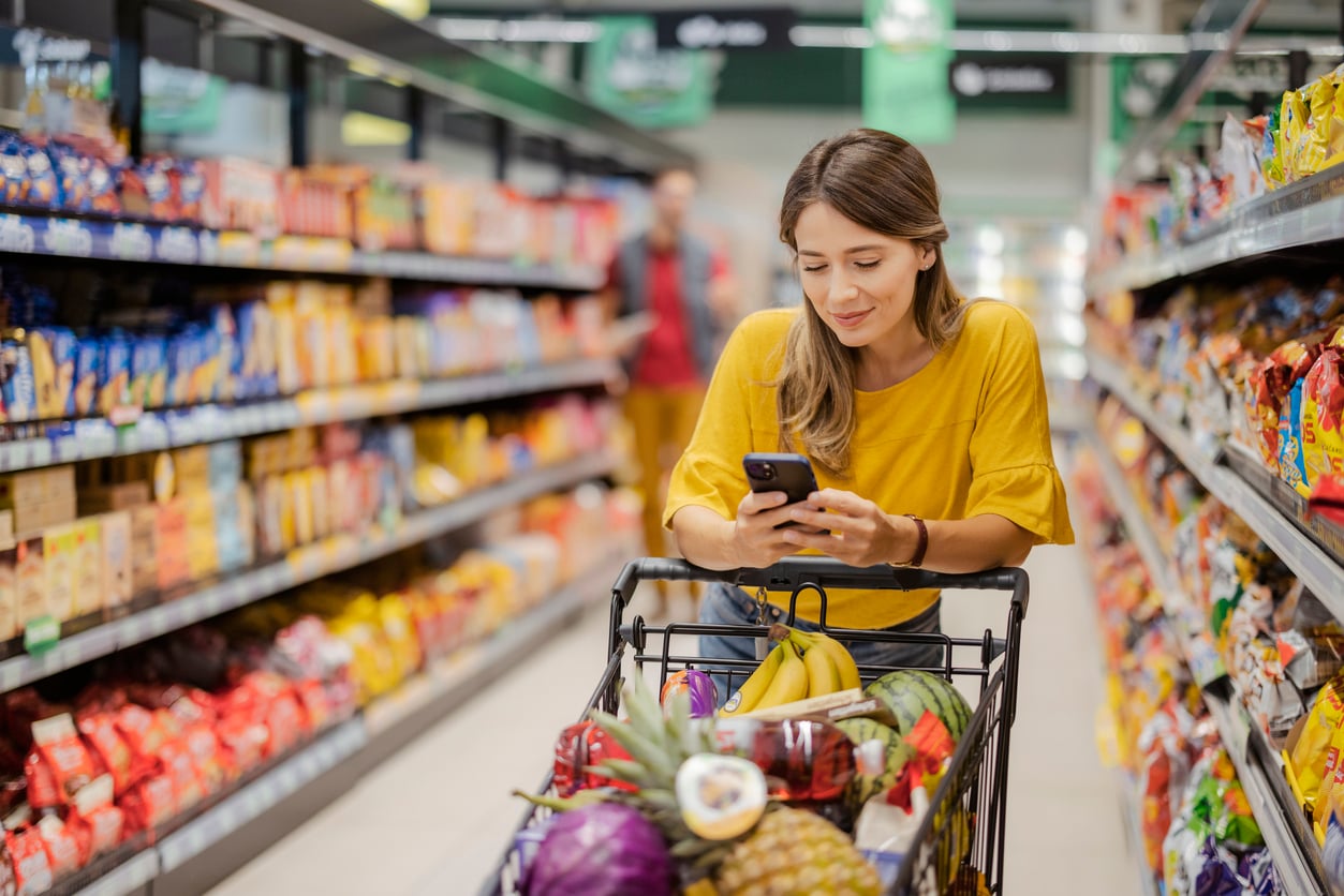 A woman in a yellow shirt pushes a shopping cart full of groceries and looks at her smartphone while standing in a supermarket aisle, thoughtfully managing her grocery habits.