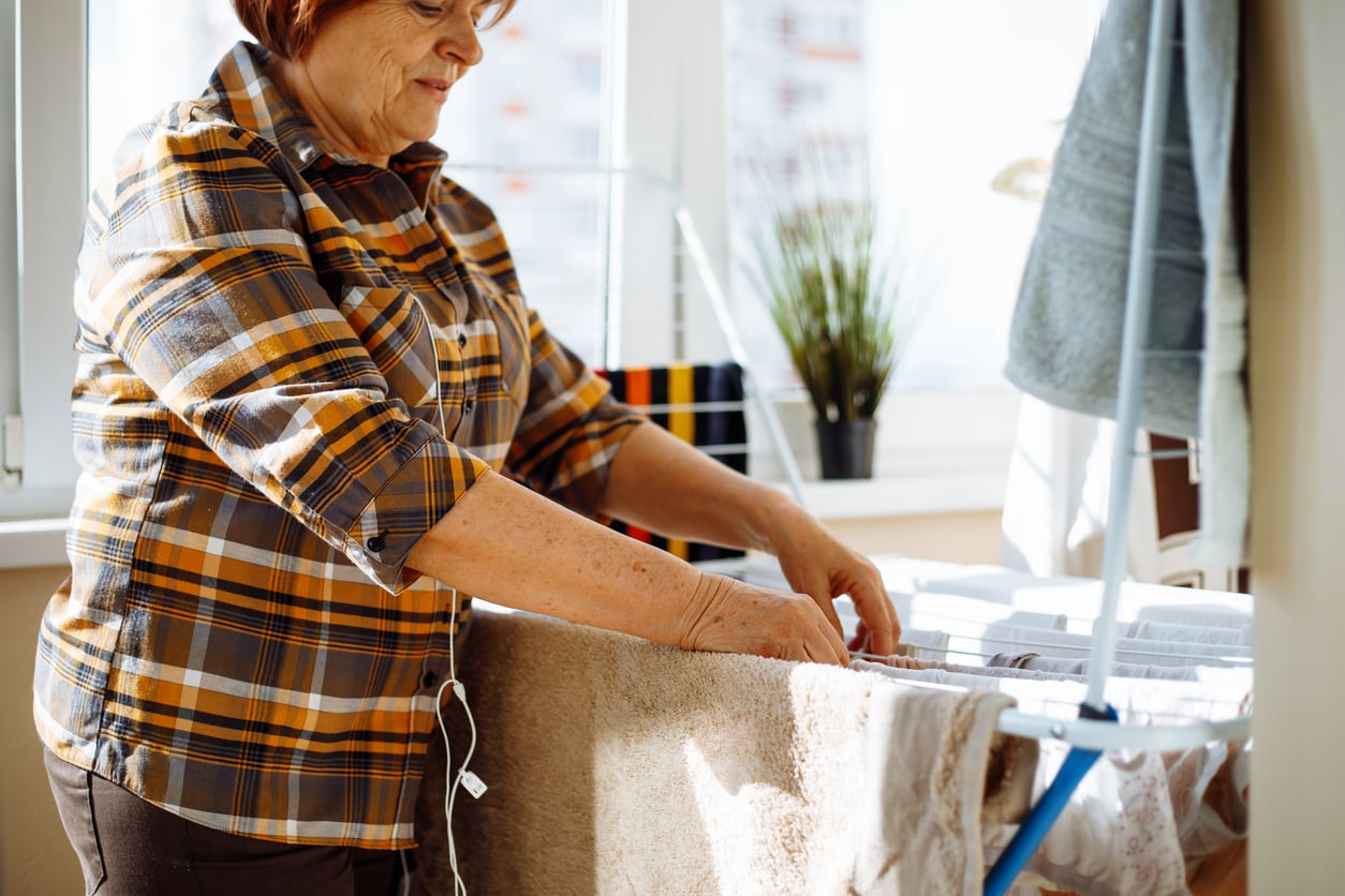 An older woman in a plaid shirt hangs freshly washed laundry on a drying rack indoors. Sunlight streams through the window, where a potted plant sits on the sill.