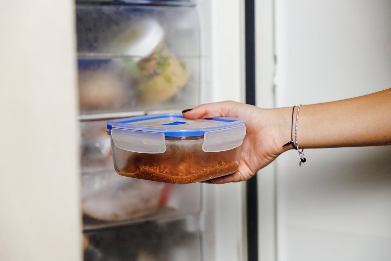 A hand is placing or removing a plastic container with a blue lid, filled with food, into a freezer with several other containers visible inside.