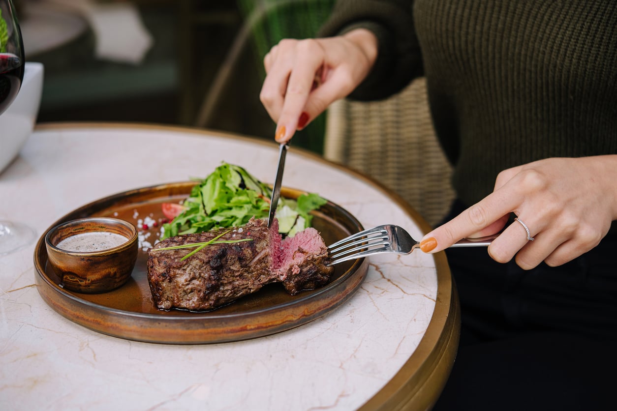 A person cuts into a medium-rare steak on a plate with greens and sauce, sitting at a marble table.