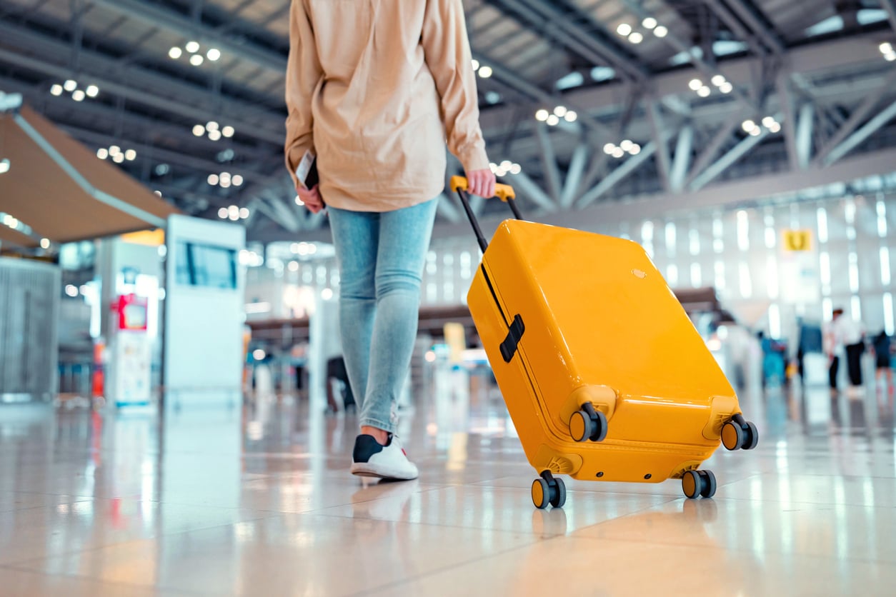 Person in jeans and a beige shirt pulls a bright yellow suitcase through a spacious, modern airport terminal with a high ceiling and large windows.