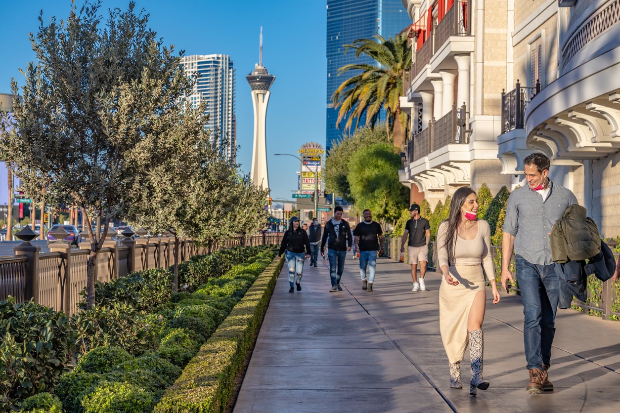 People walk along a sunny Las Vegas sidewalk with the Stratosphere Tower visible in the background, and palm trees, buildings, and green shrubs lining the path.