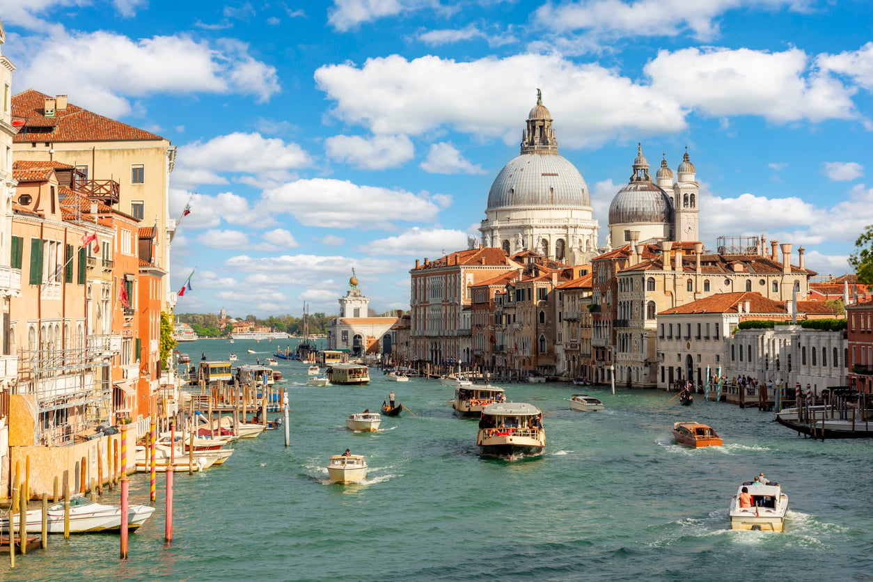 Boats travel along Venice’s Grand Canal, lined with historic buildings. The domed Basilica di Santa Maria della Salute is visible under a blue sky with scattered clouds.