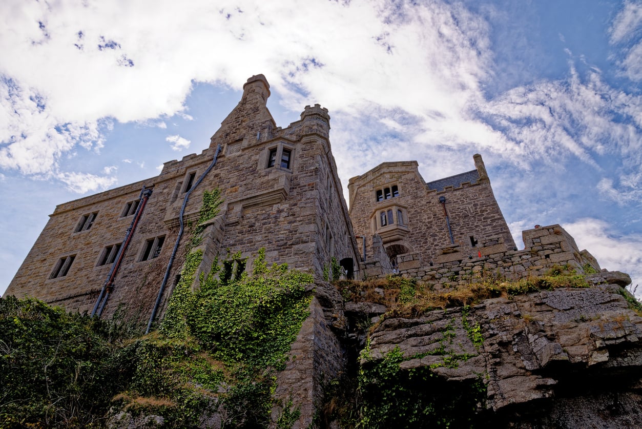 A majestic stone castle with ivy climbing its walls sits atop rocky terrain under a partly cloudy blue sky, viewed from below—an enduring symbol among castles.