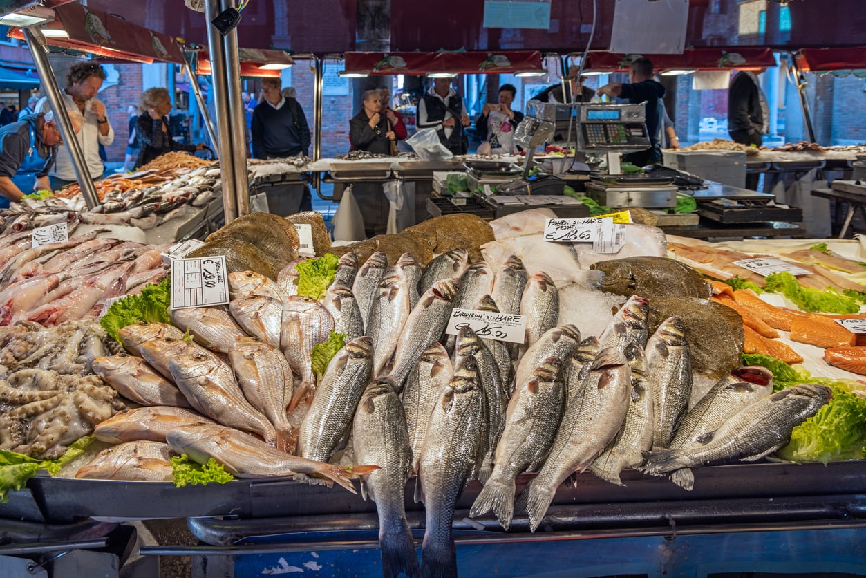 Fresh fish and seafood displayed on ice at a busy outdoor market stall, with handwritten price signs and people shopping in the background.
