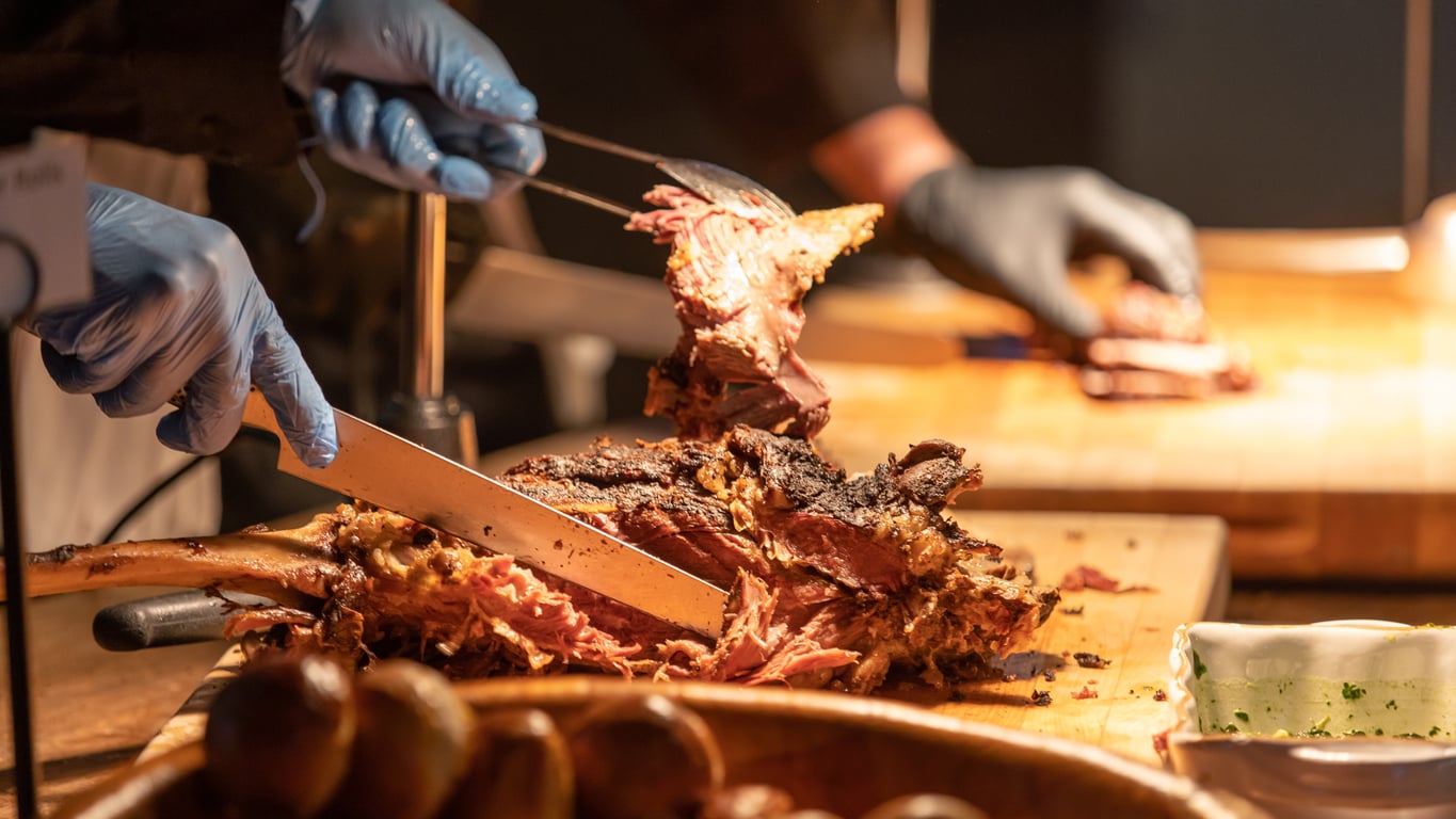 Two people wearing blue gloves carve a large piece of cooked meat on a wooden cutting board. The meat looks tender and juicy, with crispy, browned edges. In the foreground, bread rolls and a dish of sauce are visible.