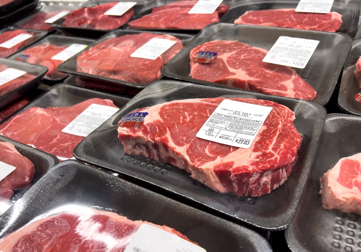 Packages of raw, marbled beef steaks in black trays with clear plastic wrap and white labels are displayed on a refrigerated shelf at a grocery store, reflecting common Grocery Habits seen among shoppers seeking fresh, quality cuts.