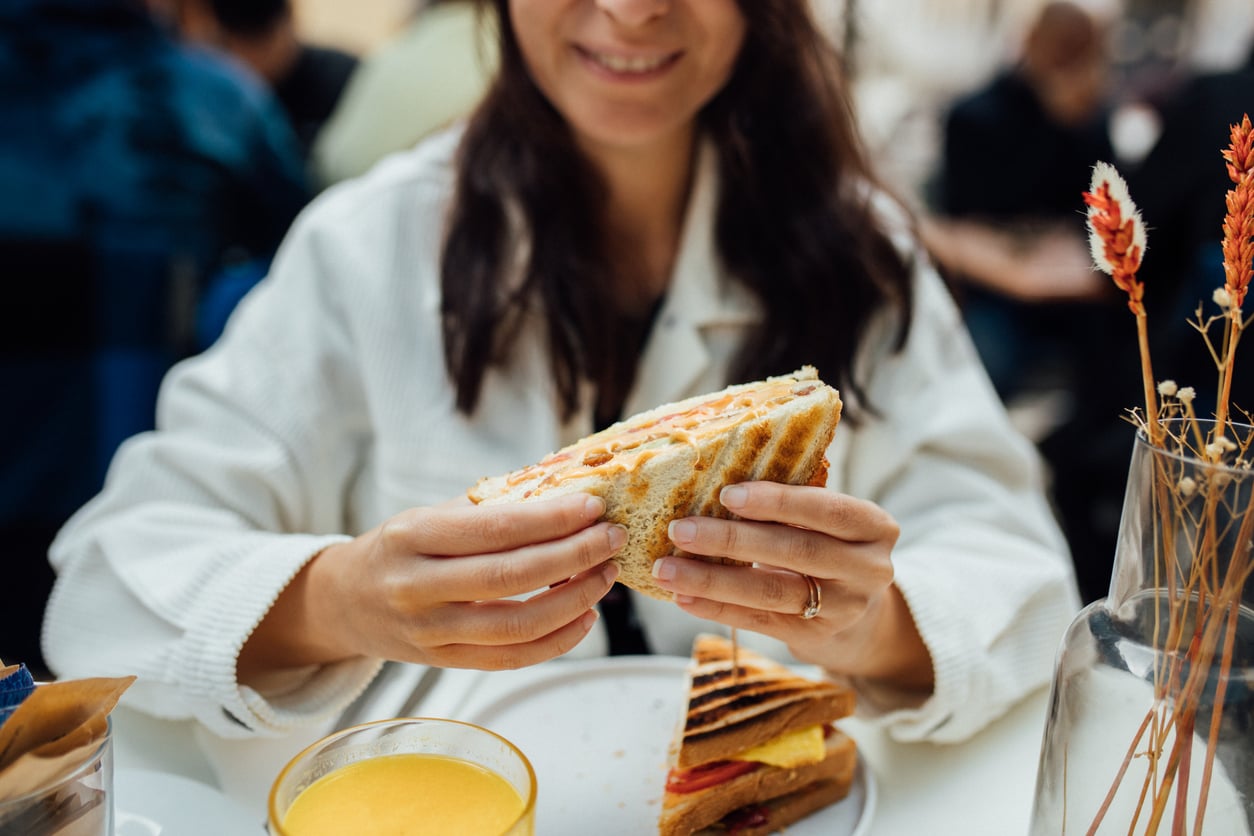 A person in a white shirt smiles while holding a sandwich at a table with another sandwich and a glass of orange juice.