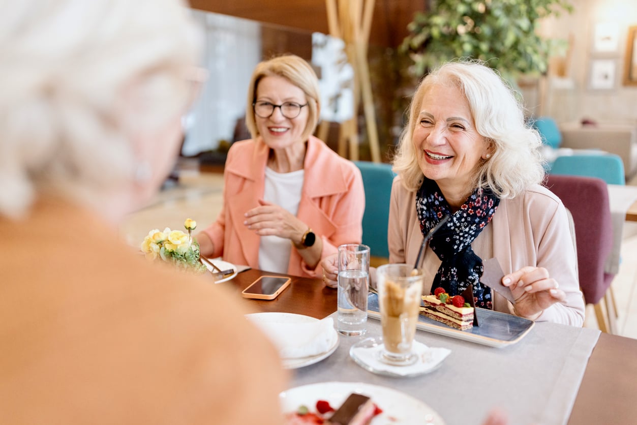 Three older women sit at a café table, smiling and enjoying dessert and drinks together. The atmosphere is bright and friendly, with natural light and cozy decor.